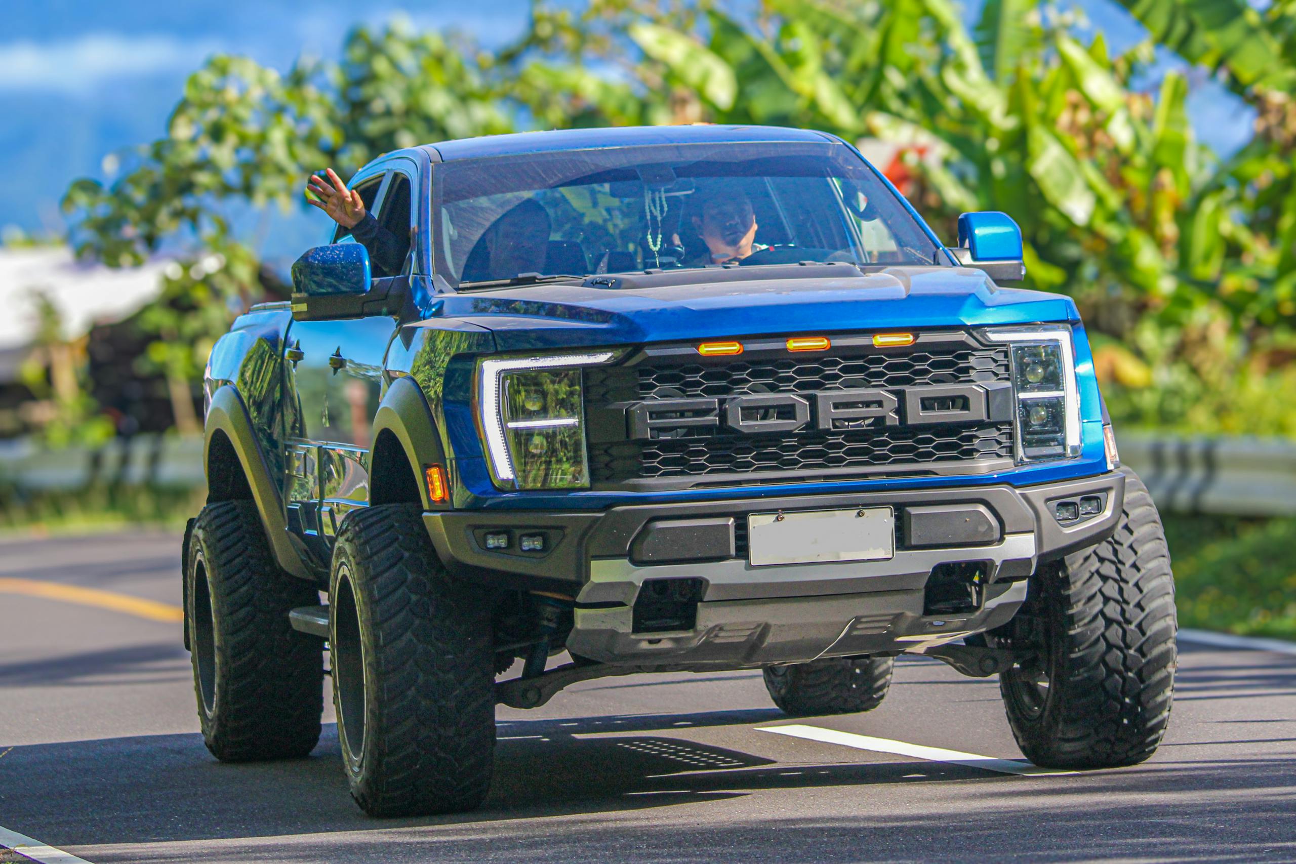 Off-road blue pick-up truck driving on a sunny scenic road with lush greenery.