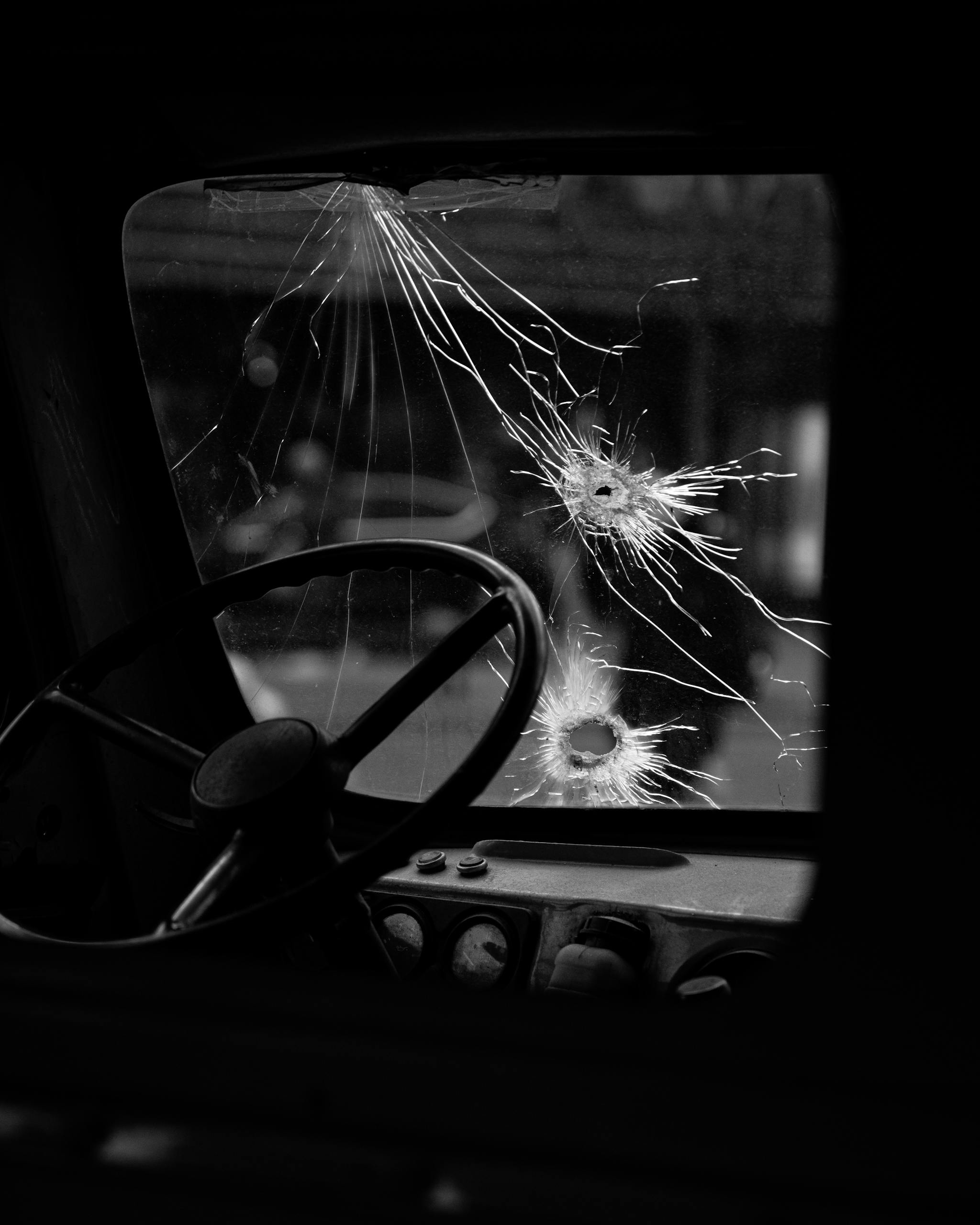Black and white photo of a broken windshield with bullet holes in an old vehicle